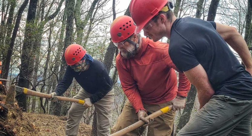 A group of people wearing hard hats use tools to work on a trail as part of a service project